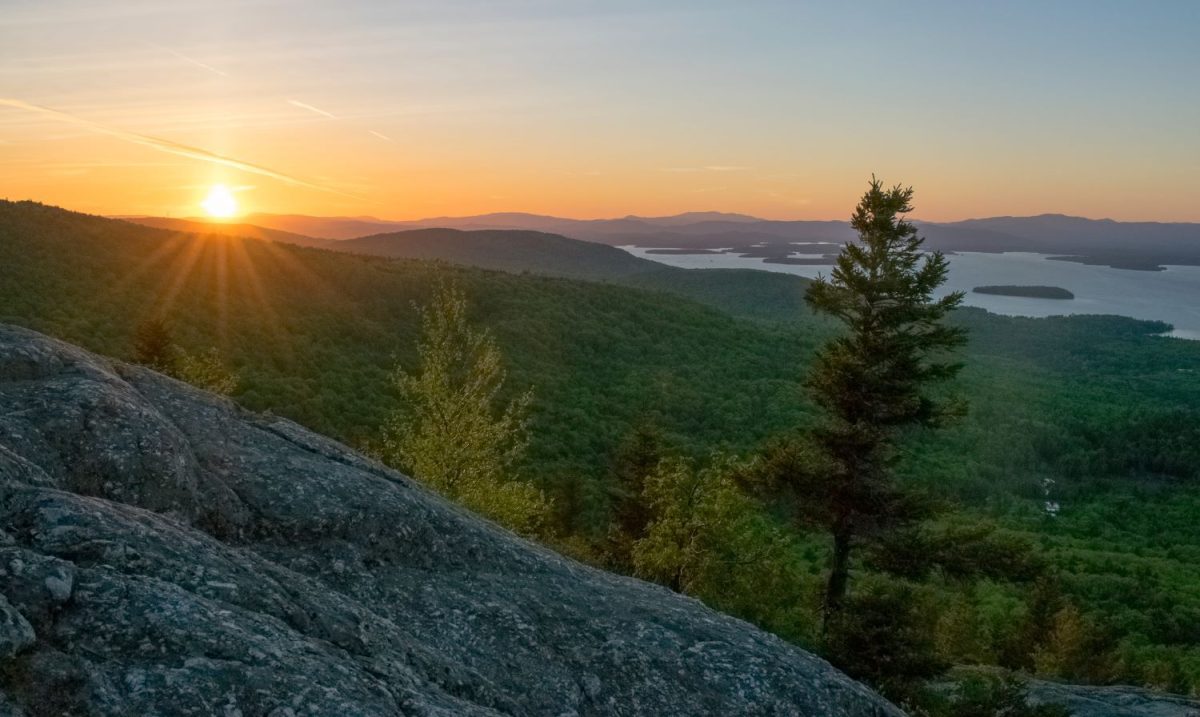 A beautiful golden sunset over the rolling green hills and distant mountain peaks surrounding Lake Winnipesaukee, showcasing the scenic hiking trails available to guests staying with Breakaway Vacation Rentals.