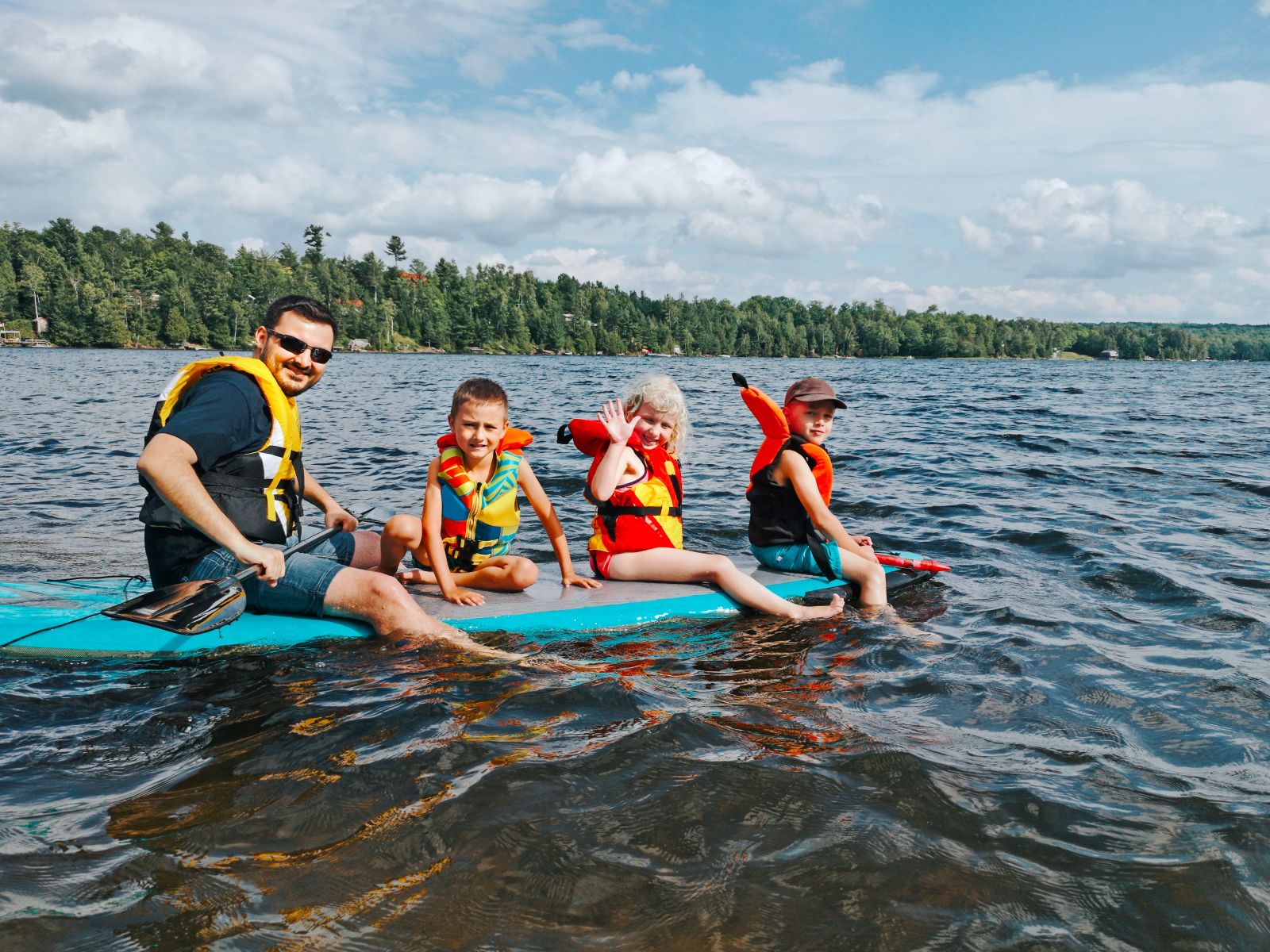 A father and his three young children wearing life jackets and smiling on a large stand-up paddleboard on the water, highlighting the family-friendly lake activities available near Breakaway Vacation Rentals in New Hampshire.
