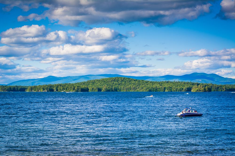 A speed boat cruising across the deep blue waters of Lake Winnipesaukee with lush islands and mountains in the background under a blue sky, illustrating the premium summer recreation options for Breakaway Vacation Rentals guests.