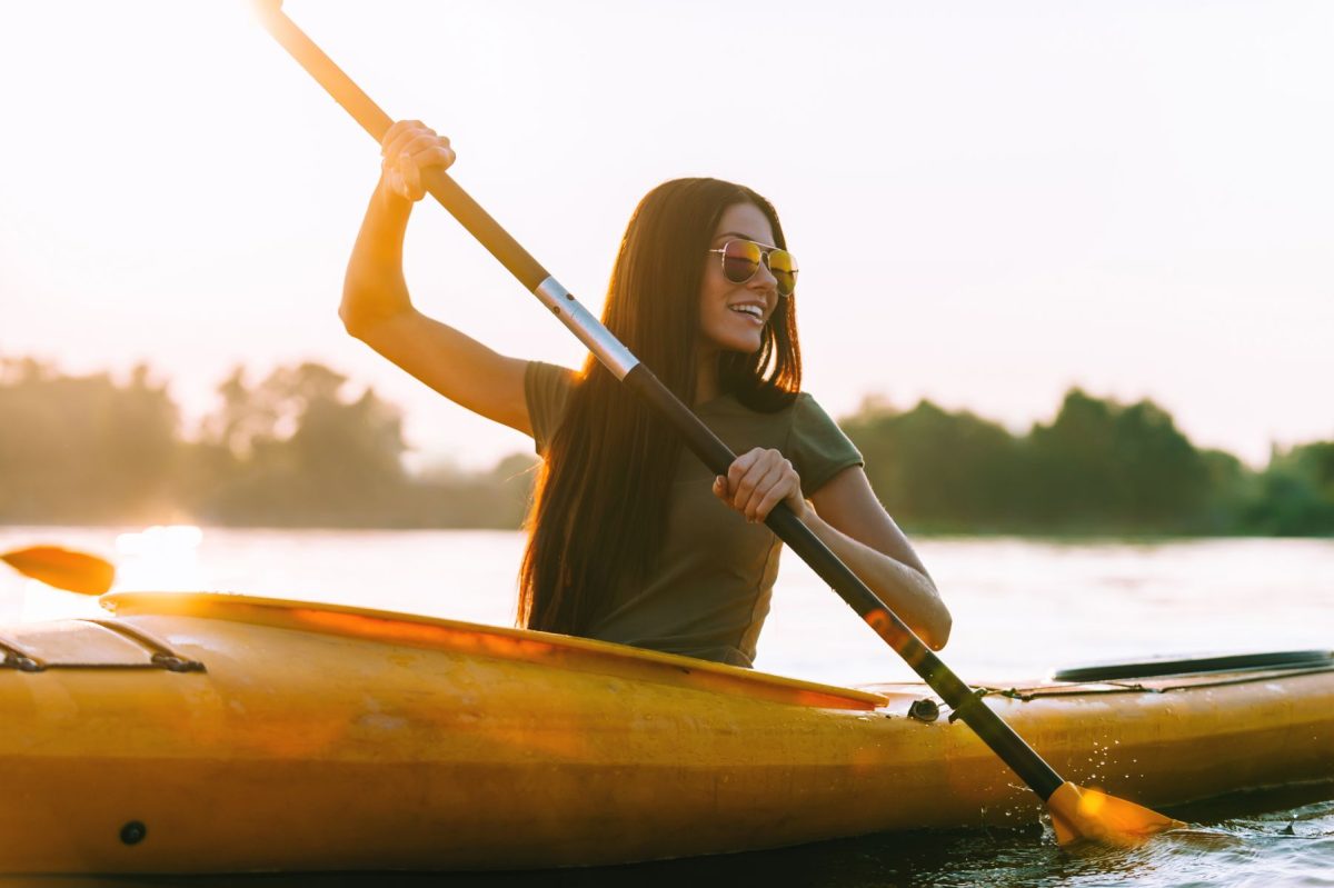 A young woman with sunglasses smiling while paddling a yellow kayak through calm lake waters at golden hour, showcasing the relaxing outdoor recreation options for guests staying with Breakaway Vacation Rentals.